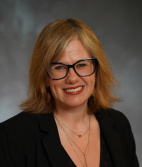 Headshot of Christie Baer, a white woman with glasses and blonde hair in a black suit jacket